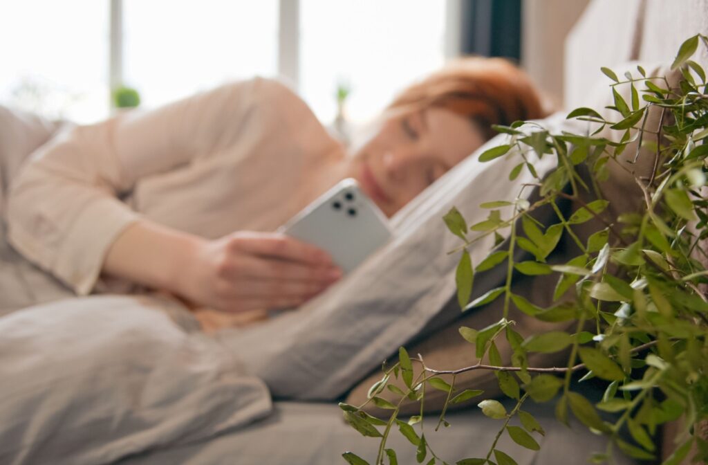 A woman lays in bed and looks at her phone after waking up in the morning.