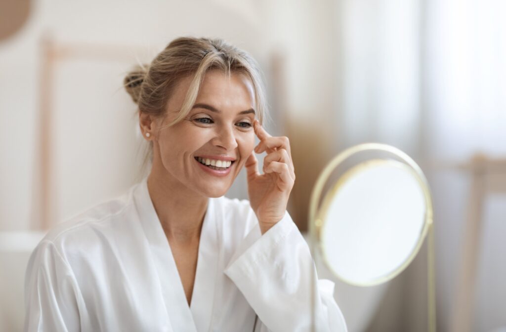 A smiling person in a white robe applies skincare near her eye while looking in a mirror.